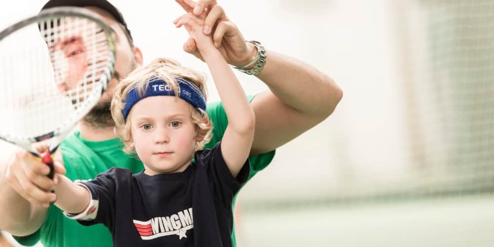 A coach helping a young child hold a tennis racket on a court.